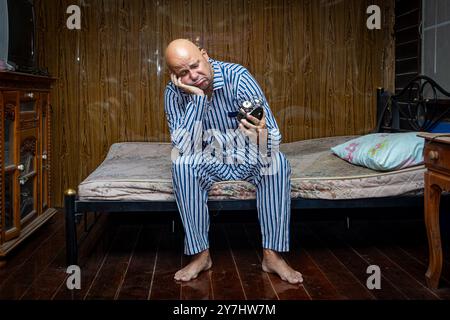 A frustrated man in pajamas sits on a mattress and looks at an alarm clock Stock Photo