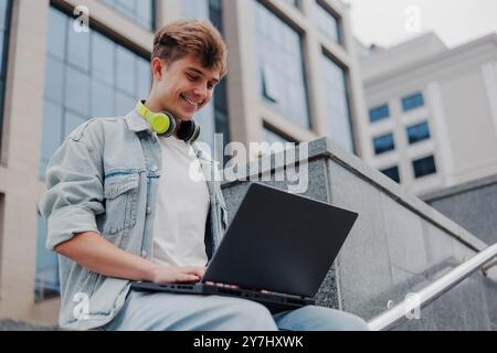 Smiling young man student or freelancer with headphones working on laptop outdoors on a background of university or corporate building. Stock Photo