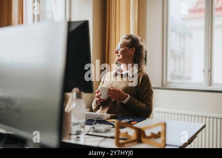 Thoughtful businesswoman holding coffee cup while sitting on chair near desk at office Stock Photo