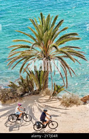 Unknown older couple moving on bicycles along Mediterranean Sea coastline with beautiful bright view of nature and palm tree. Travel, touristic place. Stock Photo
