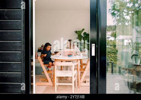Working mother assisting son doing homework sitting at home seen through sliding door Stock Photo