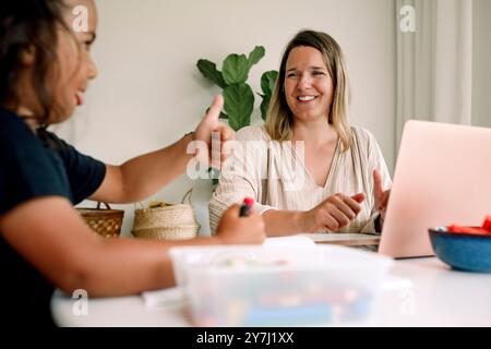 Smiling freelancer mother looking at son making thumbs up gesture while studying at home Stock Photo