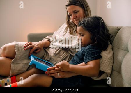 Mother and son spending leisure time watching digital tablet while sitting on sofa at home Stock Photo