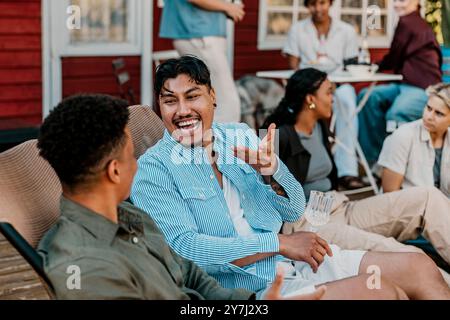 Cheerful young man enjoying drinks with male friend while sitting on chair in back yard at social party Stock Photo