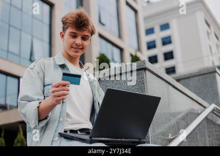 Happy young man using laptop and credit card for online shopping outdoors on a background of corporate building. Stock Photo