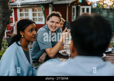 Happy young man talking with male friend while having dinner in back yard at social party Stock Photo