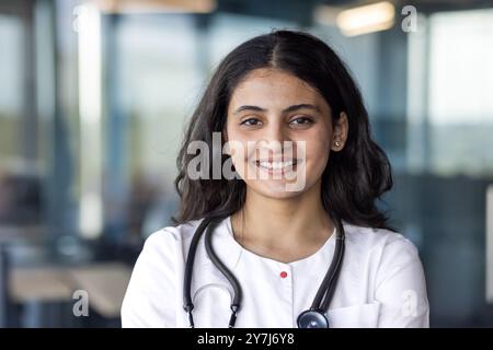 Portrait of smiling young female doctor with stethoscope. She is wearing white coat in medical office setting, conveying professionalism and confidence in healthcare role. Stock Photo