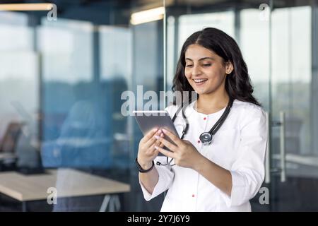 Smiling female doctor stands in modern office using digital tablet. Stethoscope around neck suggests healthcare setting. Image conveys professionalism, technology integration, and positive work Stock Photo