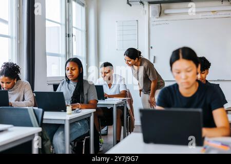 Smiling female teacher assisting student studying on laptop while sitting in classroom at school Stock Photo