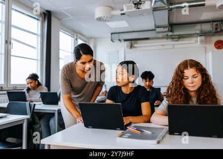 Female professor assisting teenage student sitting with laptop in classroom at junior high school Stock Photo
