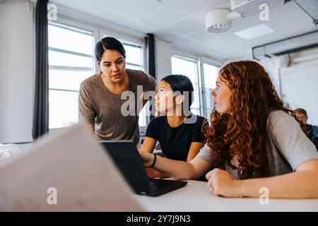 Focused teacher guiding female junior high students on laptop in classroom at school Stock Photo
