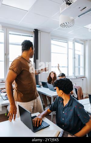 Male professor assisting students studying through laptop while sitting at desk in classroom Stock Photo
