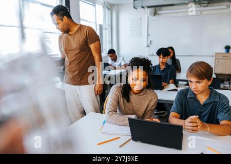 Teacher standing near male and female students doing e-learning in classroom at junior high school Stock Photo