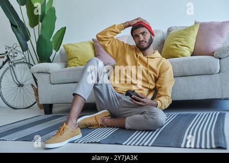 Frustrated young African man carrying joystick while sitting on the floor at home Stock Photo