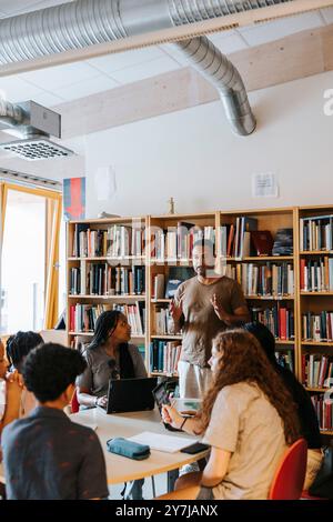 Male librarian explaining junior high students sitting near table in library at school Stock Photo