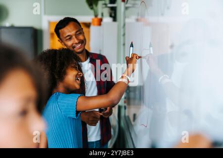 Happy girl writing on whiteboard with felt tip pen while standing near male teacher in classroom at school Stock Photo