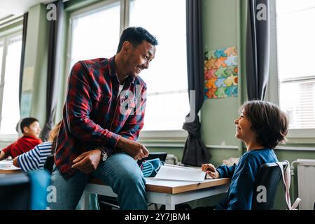 Smiling male professor sitting on desk and talking with male student sitting in classroom at elementary school Stock Photo