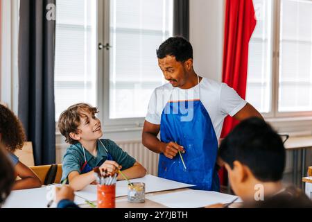 Male teacher assisting pupil painting while sitting in art class at elementary school Stock Photo