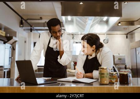 Female chef leaning on counter while discussing with male colleague in commercial kitchen Stock Photo