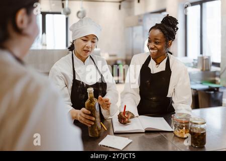 Happy young chef taking inventory with colleague holding bottle in commercial kitchen at restaurant Stock Photo