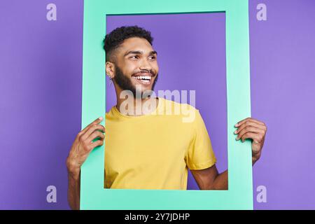 Cheerful African man looking through a picture frame while standing against purple background Stock Photo