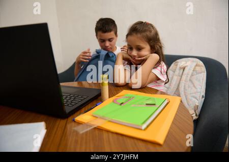 Two children sitting at a table with a laptop and school supplies, focused on learning and doing homework together. Stock Photo