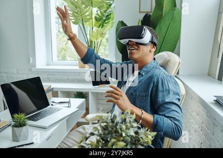 Joyful mixed race man wearing virtual reality glasses and gesturing while sitting at the desk Stock Photo
