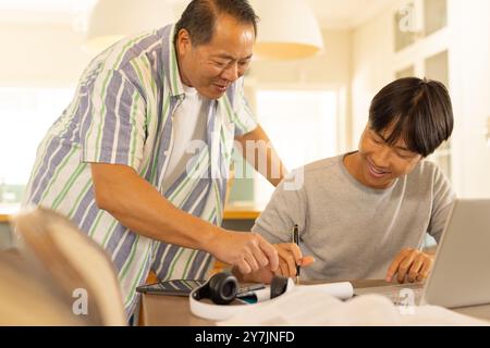Asian father helping son with homework, using laptop and writing in notebook Stock Photo