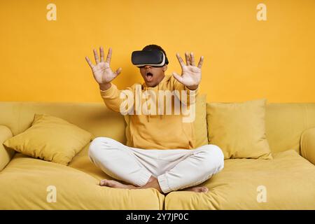 Shocked young man in virtual reality glasses relaxing on the couch with yellow background Stock Photo