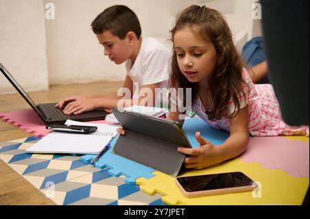Two children are engaged in using a tablet and laptop for educational and entertainment purposes, fostering digital learning at home. Stock Photo