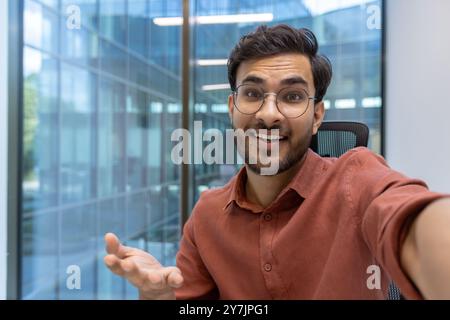 Smiling man in glasses engaged in video call using phone while sitting in modern office. Displaying friendly expression, hand gesture and casual shirt in professional setting Stock Photo