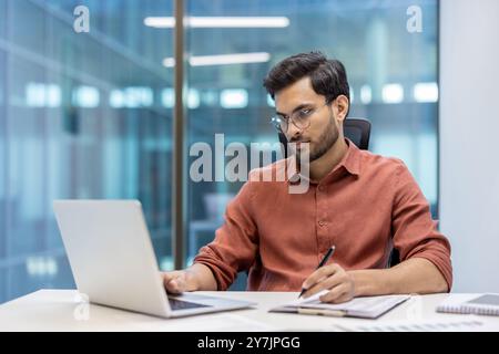 Confident man wearing glasses concentrating on laptop while taking notes. Engaged in serious paperwork, using digital tools for efficient work. Modern office environment enhances productivity focus. Stock Photo