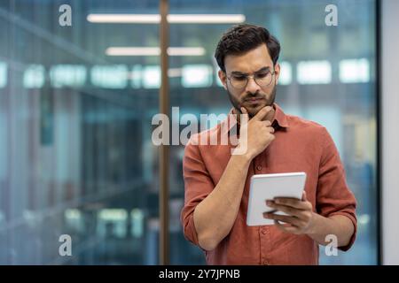 Serious programmer focused on tablet screen in modern office. Individual wearing glasses and casual attire analyzing information to solve problems or develop software. Stock Photo