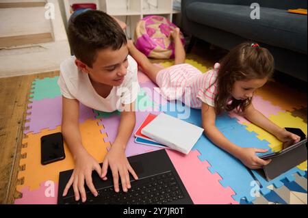 A boy using a laptop and a girl with a tablet, focusing on digital learning in a cozy home setting. Stock Photo