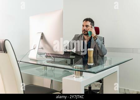 Middle-aged professional man in stylish office setting, using smartphone while working on computer. Modern workplace with glass desk, depicting multit Stock Photo