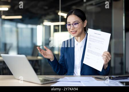 Confident Asian businesswoman leads virtual meeting. Engaging tone, holding document, gestures towards laptop screen. Demonstrates modern business practices, effective communication Stock Photo