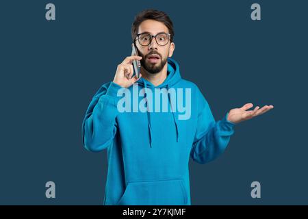 Young man in a blue hoodie talking on the phone with a surprised expression Stock Photo
