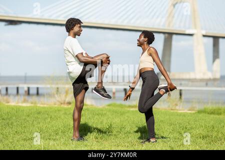 Two friends stretching together at a park near a river during daytime Stock Photo