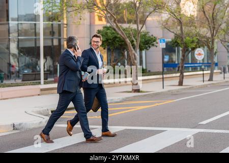 Two attractive confident hispanic businessmen in suits walking outdoors at the city streets. Two businessmen walking and talking at urban city. Middle Stock Photo