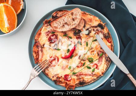 Italian frittata or omelette with tomatoes, cheese and toasted toast on a blue plate with cutlery and napkin on a white table with a bowl of oranges. Stock Photo