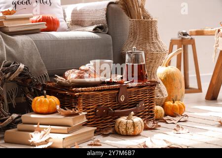Autumn composition with teapot, cup of hot beverage, tasty buns and pumpkins on wicker basket in living room Stock Photo
