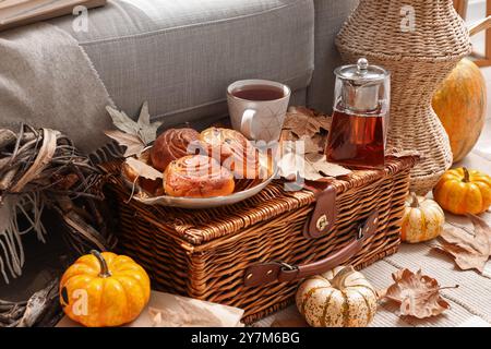 Autumn composition with teapot, cup of hot beverage, tasty buns and pumpkins on wicker basket in living room Stock Photo