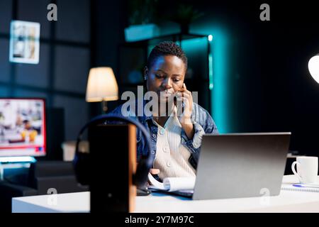 African american female freelancer utilizing laptop in home office while chatting on mobile device with customer. Black woman working remotely, reviewing documents and making phone calls. Stock Photo