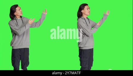 Asian guy doing advertisement on camera, pointing at something next to him for new marketing ad. Young adult smiling and working on promotional slogan, standing over greenscreen backdrop. Stock Photo