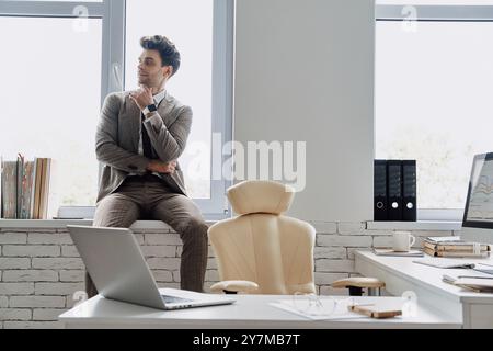 Thoughtful man in formalwear holding hand on chin while sitting on the window sill in office Stock Photo