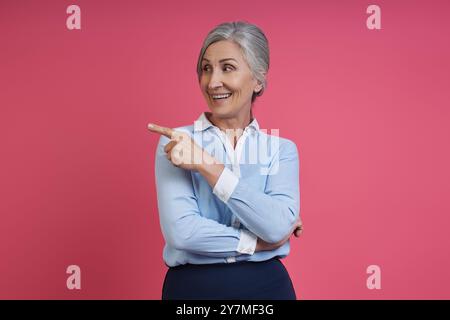 Confident senior woman pointing away and smiling while standing against pink background Stock Photo