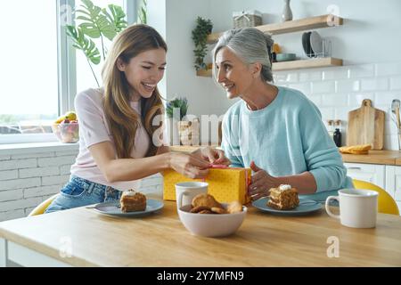 Happy young woman receiving a gift box from her mother while both sitting at the kitchen Stock Photo