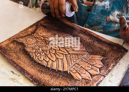 Close-up of an artisan applying varnish to a copper sheet engraved with a detailed owl motif, using an aging technique to preserve the vintage, weathe Stock Photo