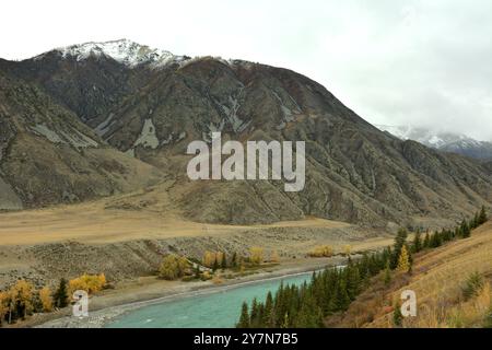 A look at a beautiful turquoise river with pines and larches on its banks flowing through an autumn valley near a high mountain with a snow-covered pe Stock Photo