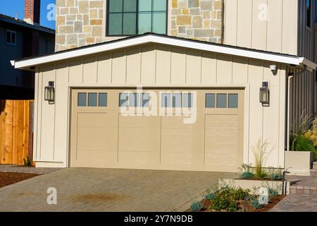 A modern beige garage door with four glass panels is part of a contemporary home, featuring a stone and siding exterior. The driveway is paved with br Stock Photo